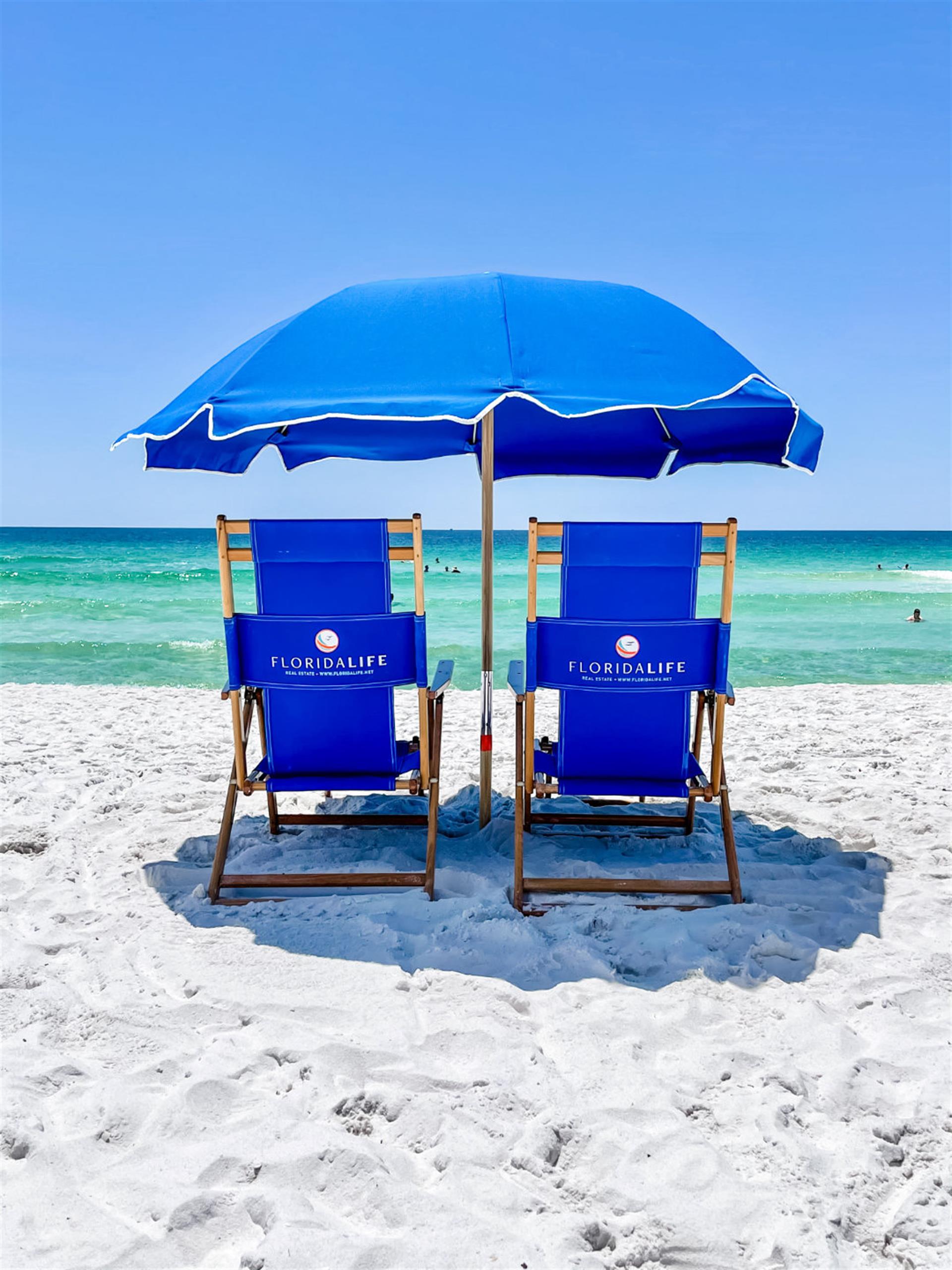 Two blue beach chairs in the sand facing the ocean with a blue umbrella shading them