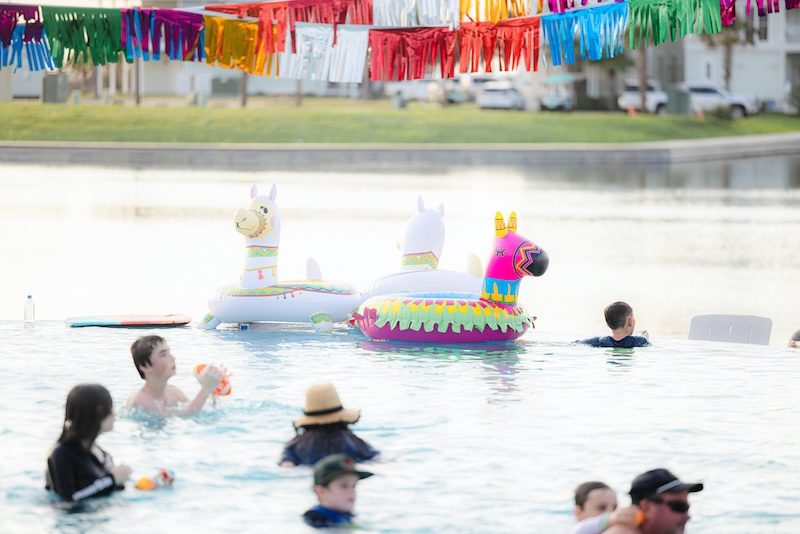 Families swimming with colorful llama floats under vibrant papel picado banners at the Fiesta by the Shore Cinco de Mayo pool party at Cinnamon Shore in Port Aransas, Texas