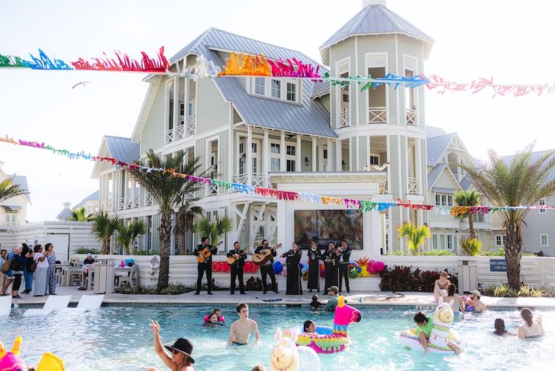 Mariachi band performing poolside at the Fiesta by the Shore Cinco de Mayo celebration at Cinnamon Shore in Port Aransas, Texas, with colorful papel picado banners and families enjoying the resort-style pool
