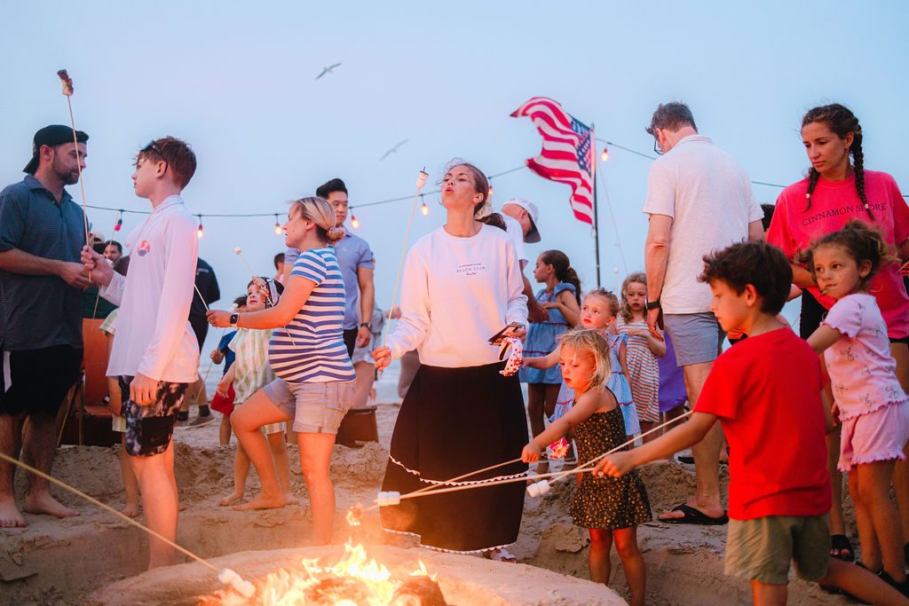 Families and children roasting marshmallows over a beach bonfire with the American flag and string lights at the Cinnamon Shore Memorial Day celebration in Port Aransas, Texas