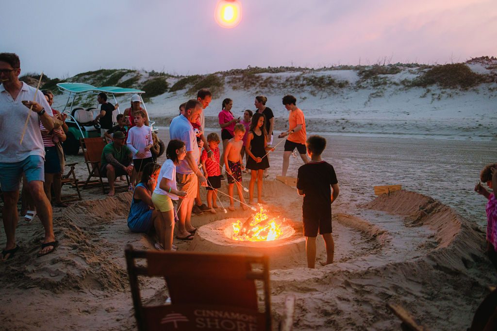 Kids and families gathered around a sand fire pit roasting marshmallows at sunset at Cinnamon Shore's Memorial Day event in Port Aransas, Texas