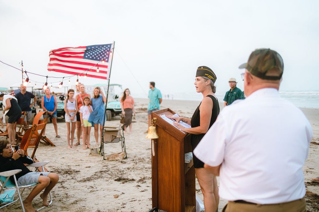 Veteran speaking at a podium with a ceremonial bell on the beach during the Cinnamon Shore Memorial Day flag ceremony in Port Aransas, Texas