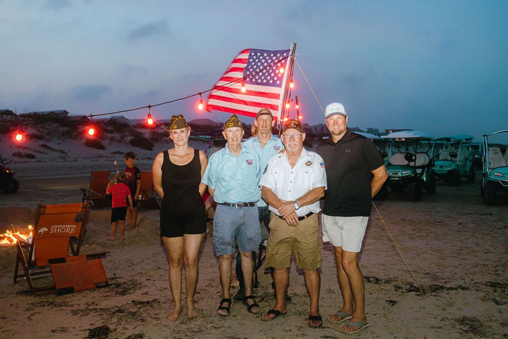 Group photo of veterans and community members standing together on the beach with the American flag and string lights at dusk during the Cinnamon Shore Memorial Day celebration