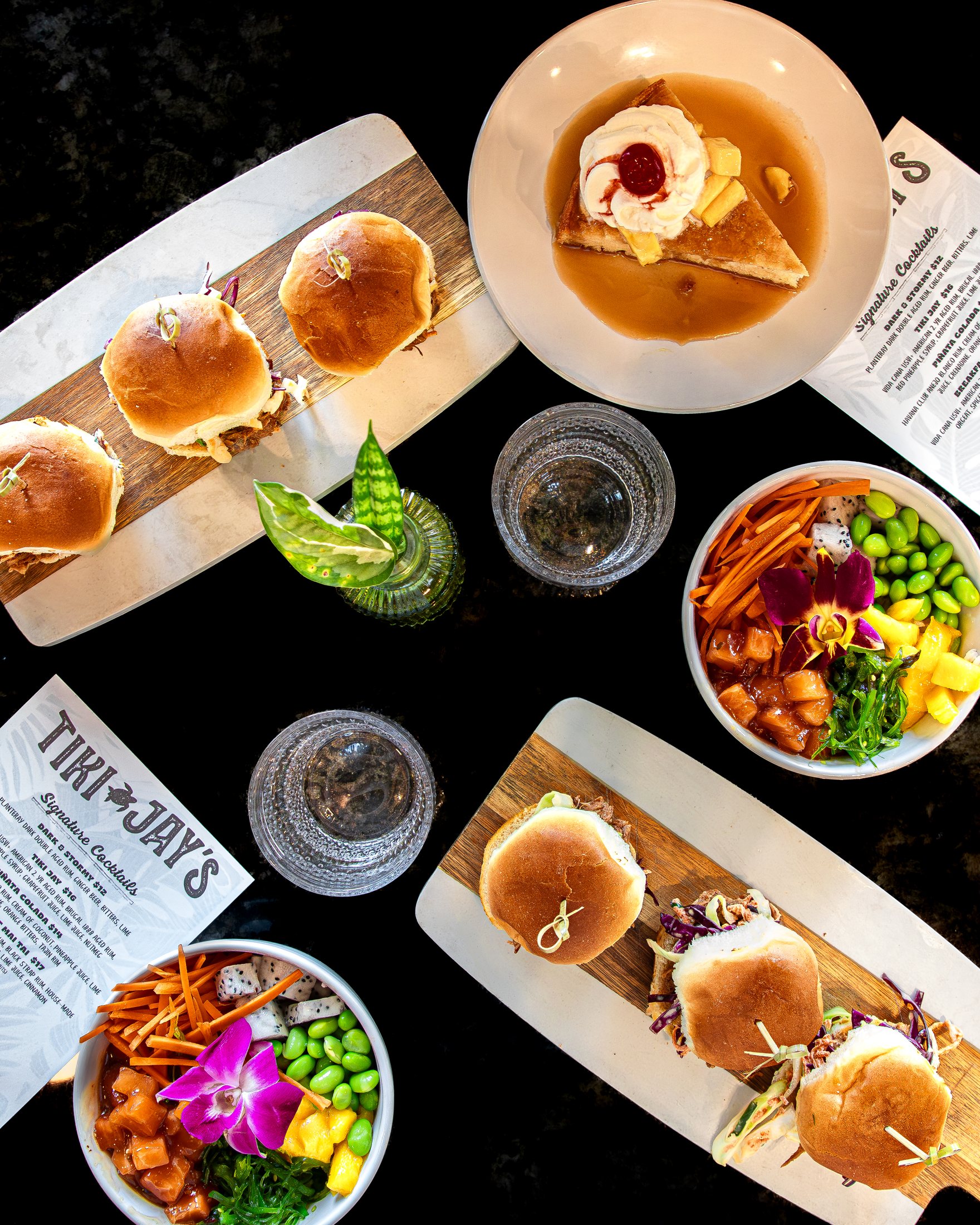 Overhead spread of Tiki Jay's dishes with a poke bowl, sliders, small plates, and drinks at Cinnamon Shore