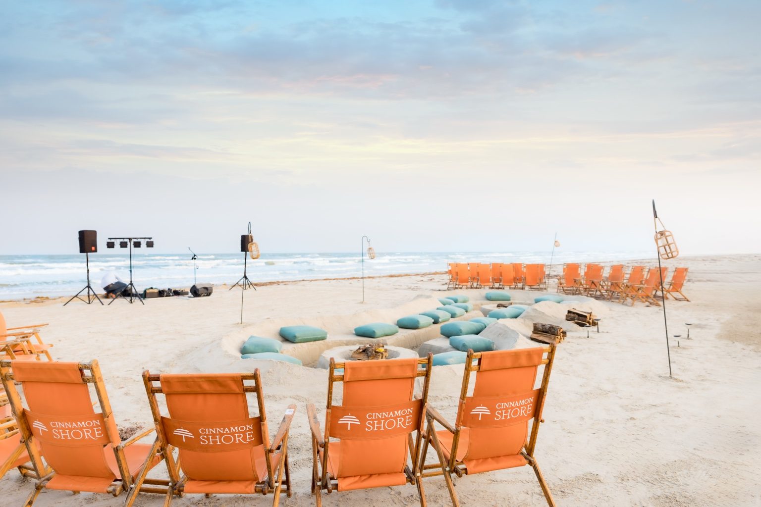 Gulf-front Cinnamon Shore bonfire setup with branded beach chairs, turquoise cushions, lanterns, and live music at sunset.