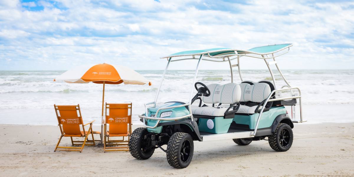 A six-seater Cinnamon Shore golf cart parked on the beach beside a branded orange umbrella and beach chairs by the Gulf.