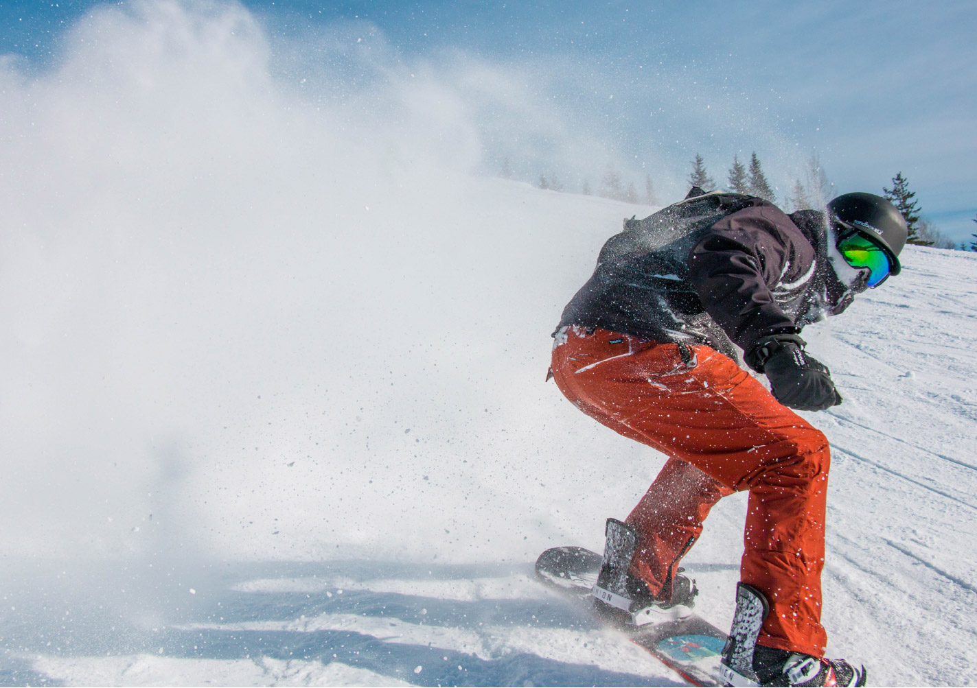 A man wearing orange ski pants, a black winter coat, and helmet snowboards down a mountain in Park City, Utah.