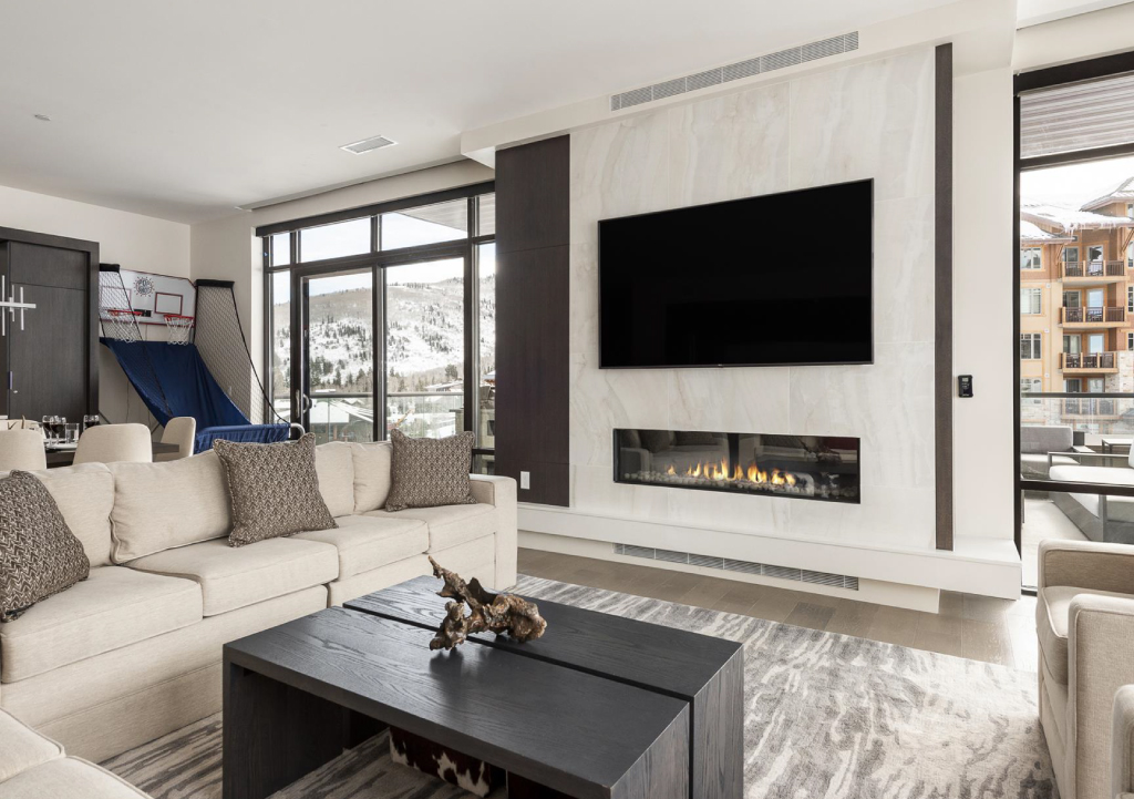 Modern living room with large TV above sleek gas fireplace and view of snowy mountains outside.