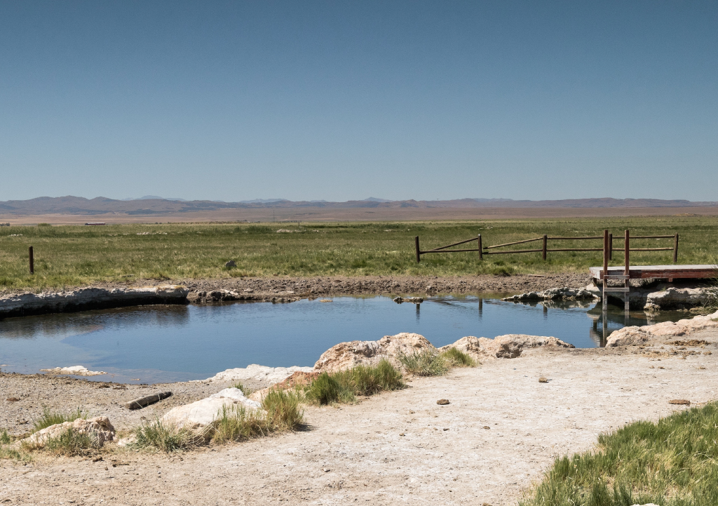 Pool of water surrounded by flat open land with mountains in the distance at Inlet Park Hot Springs in Utah.