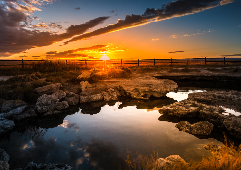 Brilliant orange sunset reflecting off a pool of water surrounded by open land at Meadow Hot Springs in Utah.