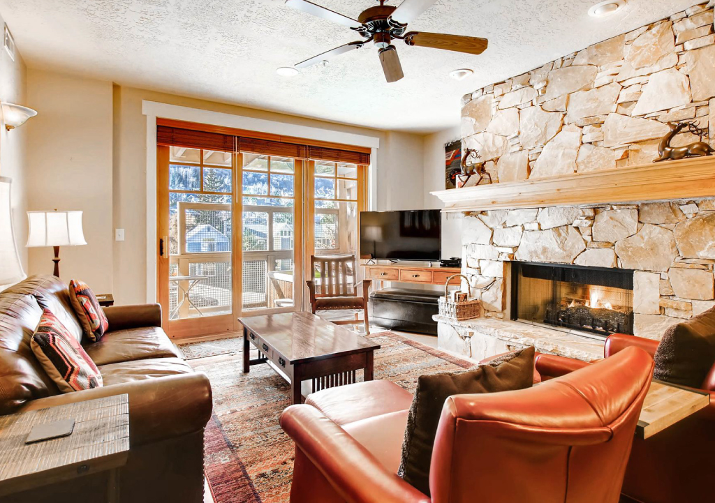 Inviting, rustic living room with large stone fireplace and TV, leading out to a balcony overlooking mountains.