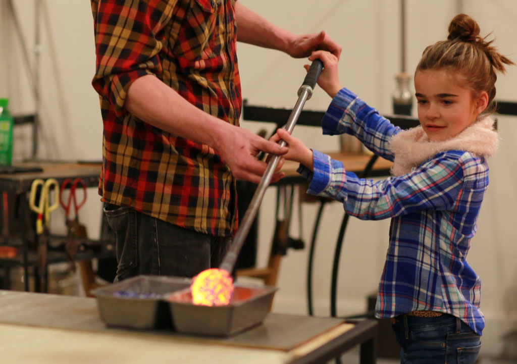 Child holding glass-blowing equipment with the assistance of an adult.