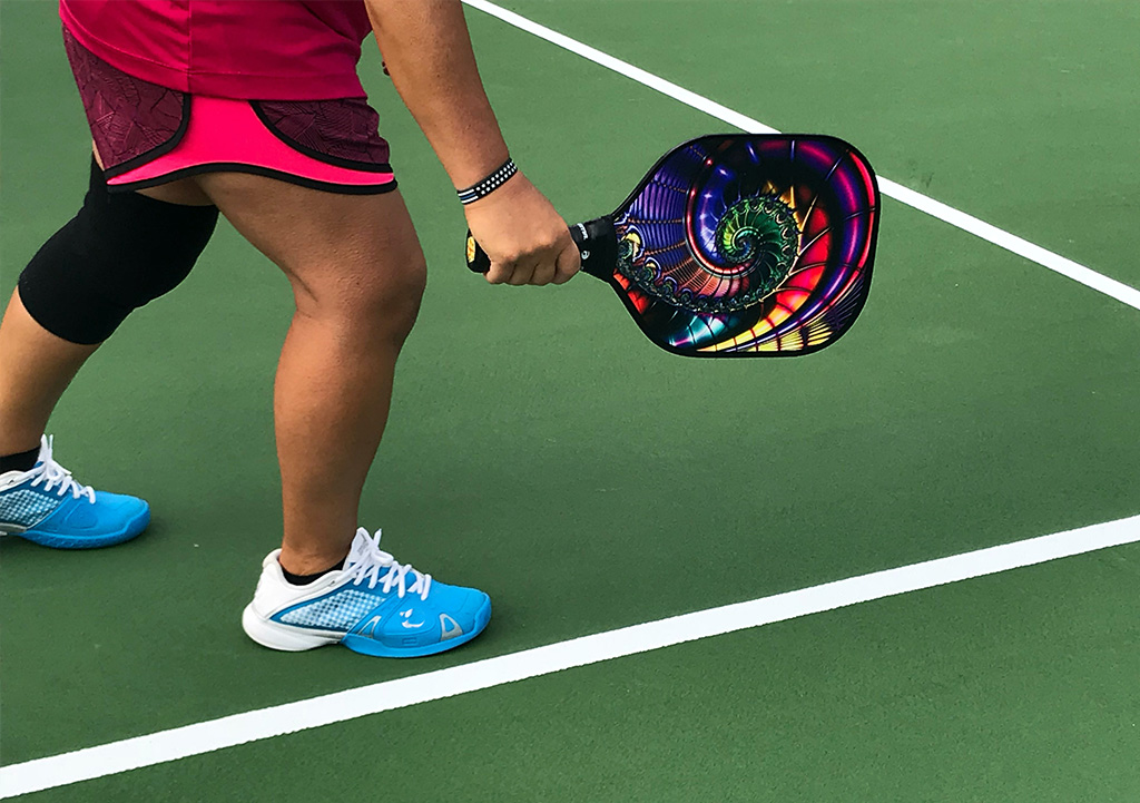 Person's legs and arm holding a pickleball racket with many colors on a pickleball court.