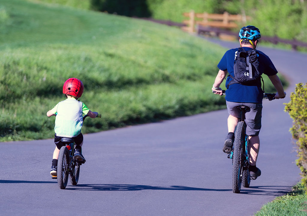 A parent and young child riding bikes next to each other along a paved trail surrounded by grass.