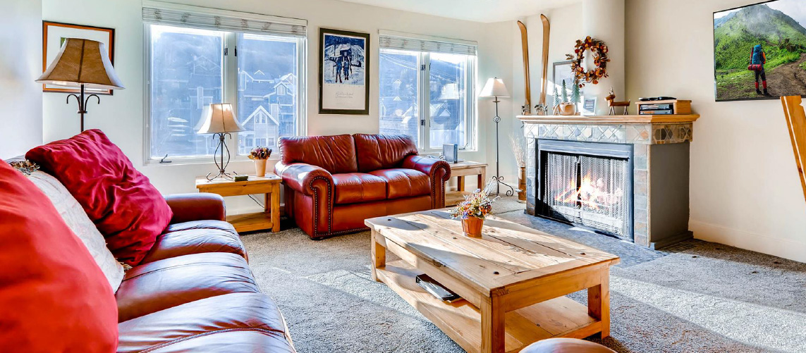 Rental condo living room with grey carpet, wood burning fireplace, red leather couched, and rustic wood tables.