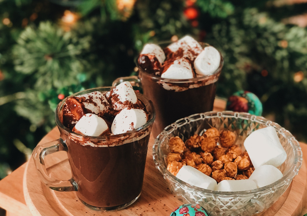 Two glass mugs of hot chocolate with marshmallows next to a bowl of popcorn and marshmallows on a small table with a Christmas tree in the background. 