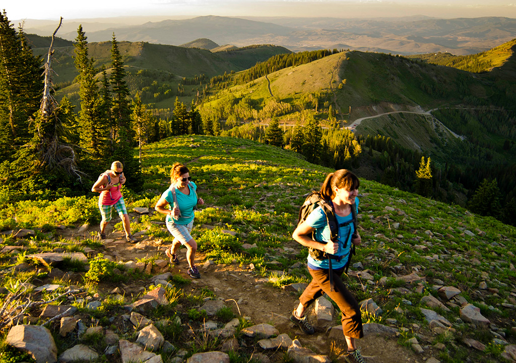 Three people hike dirt trail on green, rolling hillside at sunset.