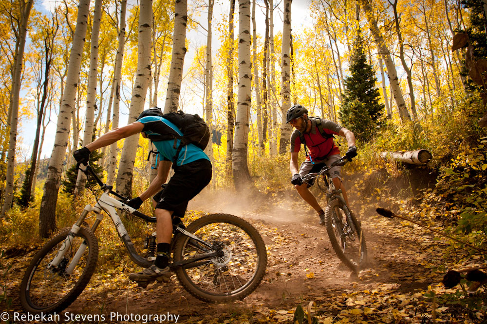 Two mountain bikers turn a sharp trail curve in a forest in the fall.