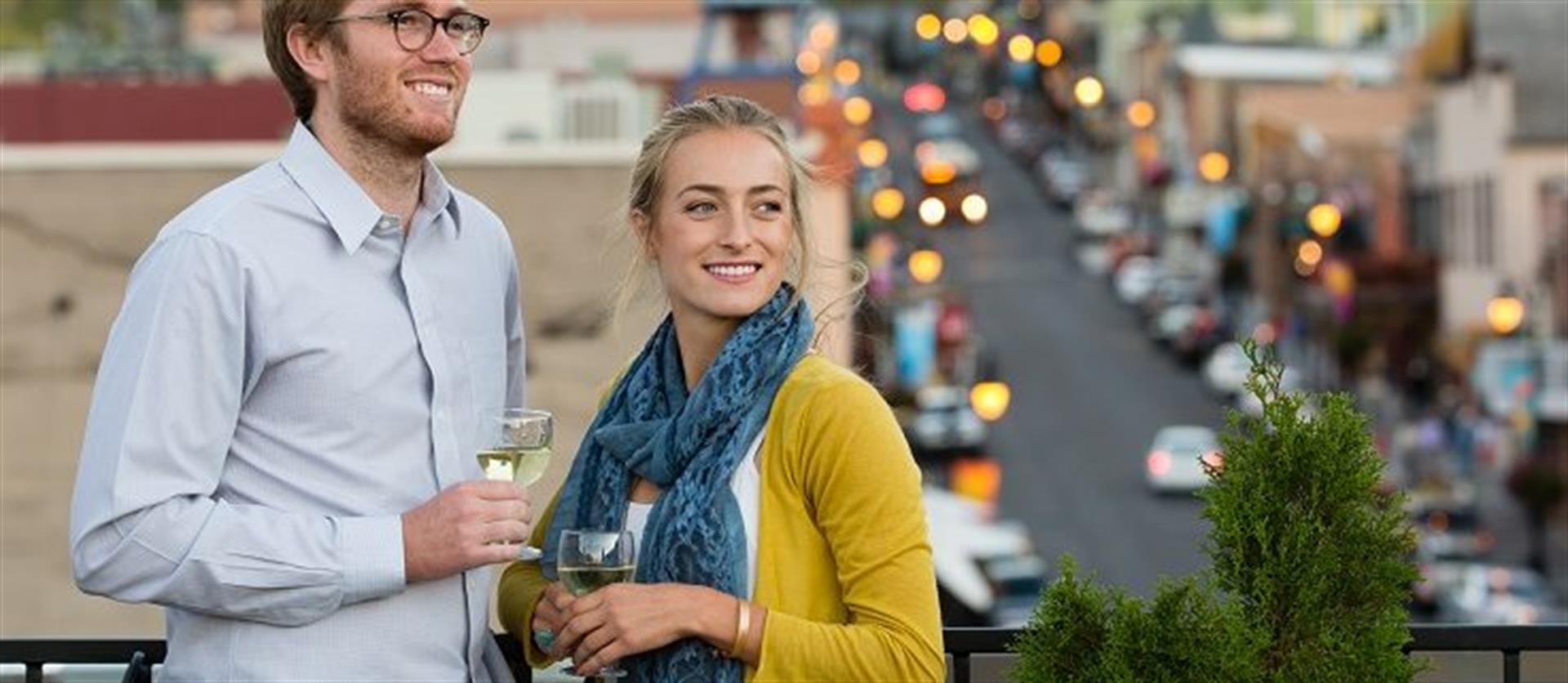 A couple wearing blue and yellow tones holding glasses of wine, standing on a balcony overlooking Main Street