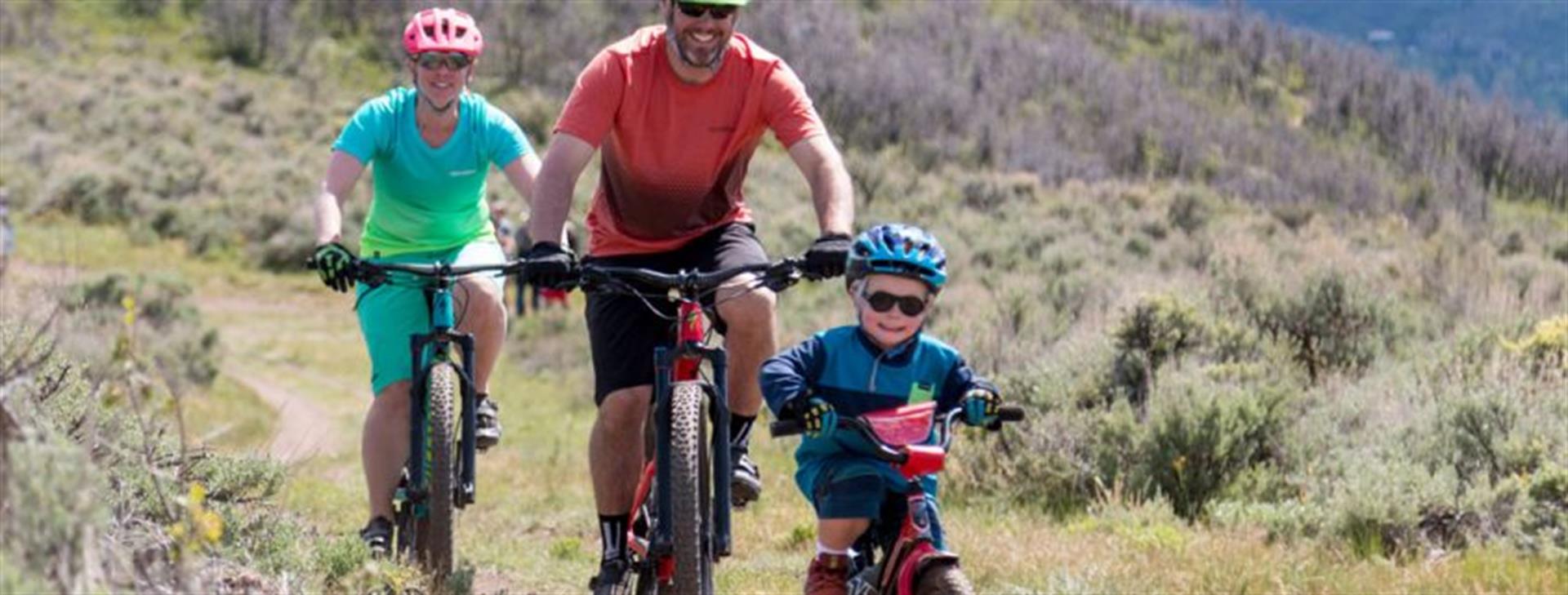 A mom, dad, and son on mountain bikes, coming down a single track through sage brush and grassland