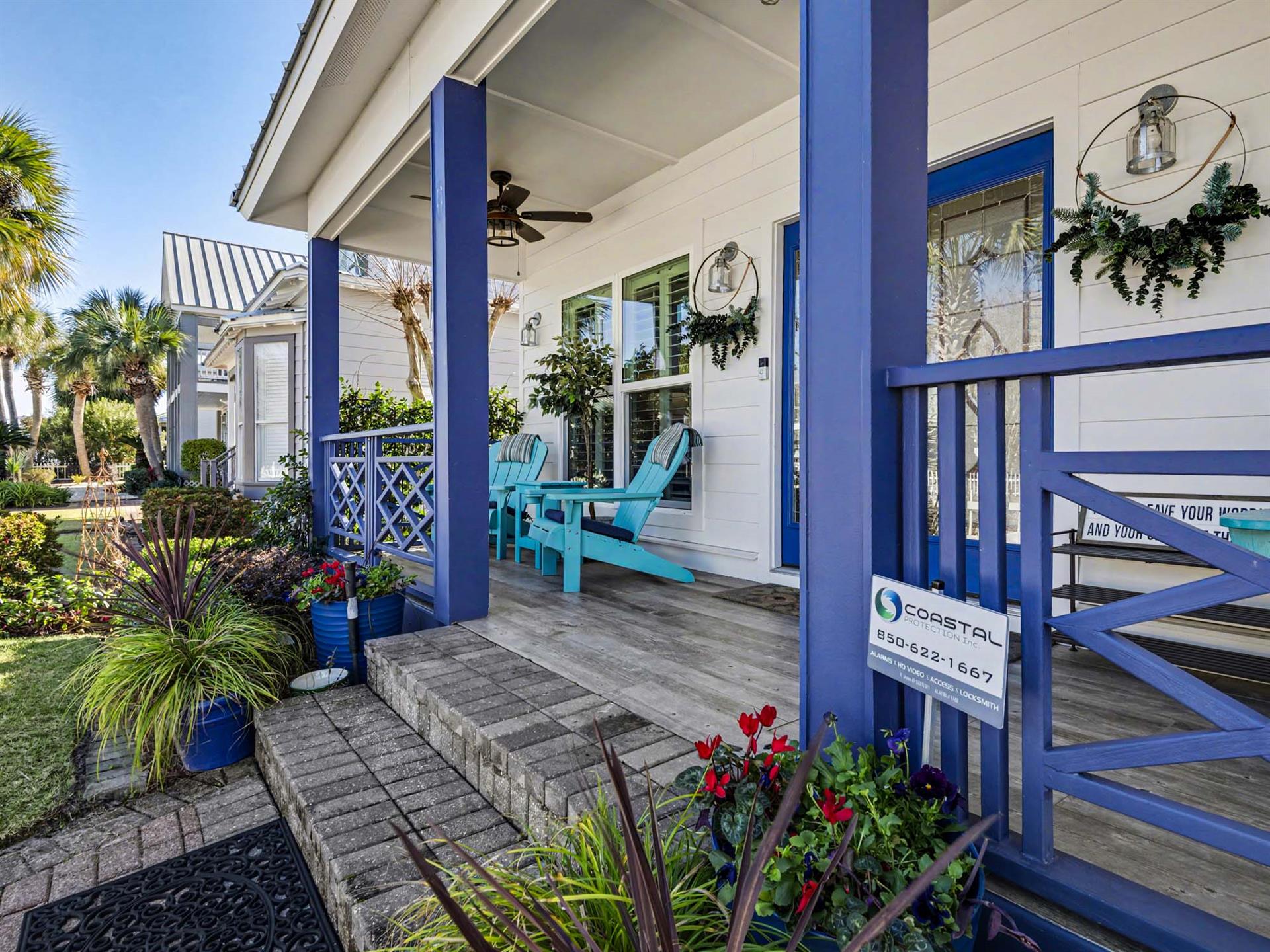 Coastal property entrance featuring tropical landscaping, turquoise Adirondack chairs, and blue accents against white siding.