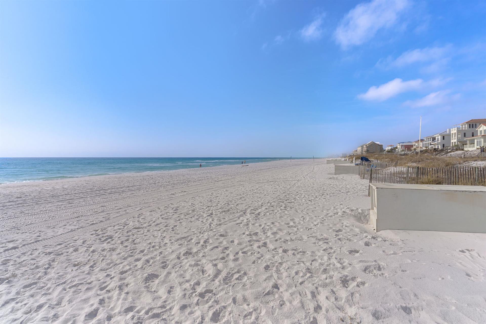 Pristine white sand beach stretches along turquoise waters with coastal homes dotting the shoreline under clear blue skies.