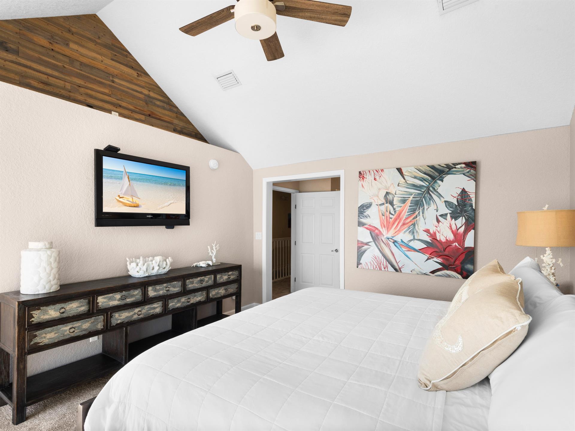 Drift into coastal dreams in the serene primary bedroom, featuring a plush king bed, tropical artwork, and rustic wood accents that create the perfect retreat.
