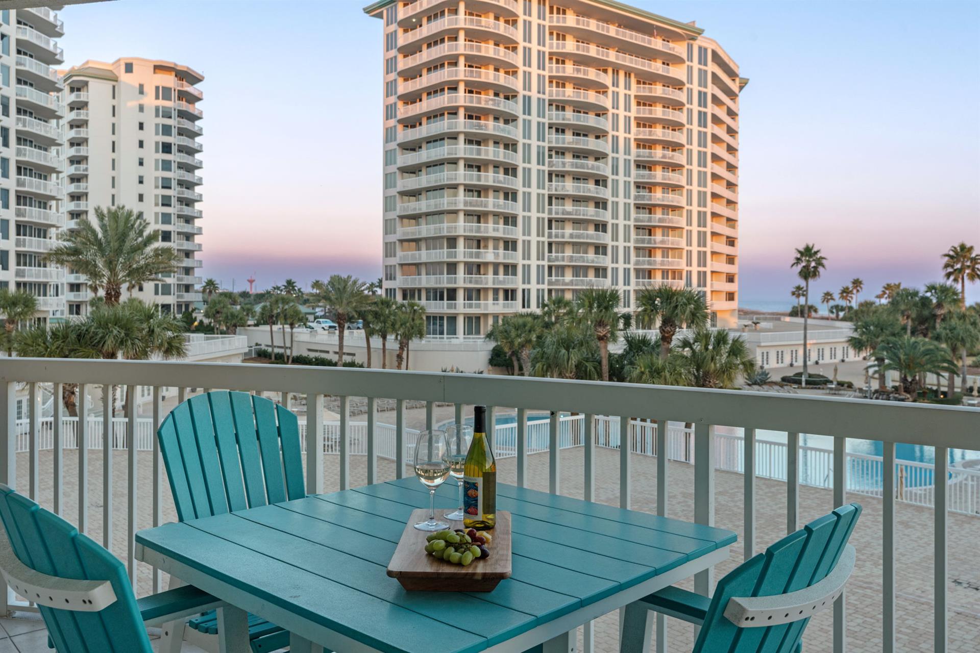 Beachfront balcony with colorful Adirondack chairs, wine setup, and views of sand, palm trees, and coastal highrises.