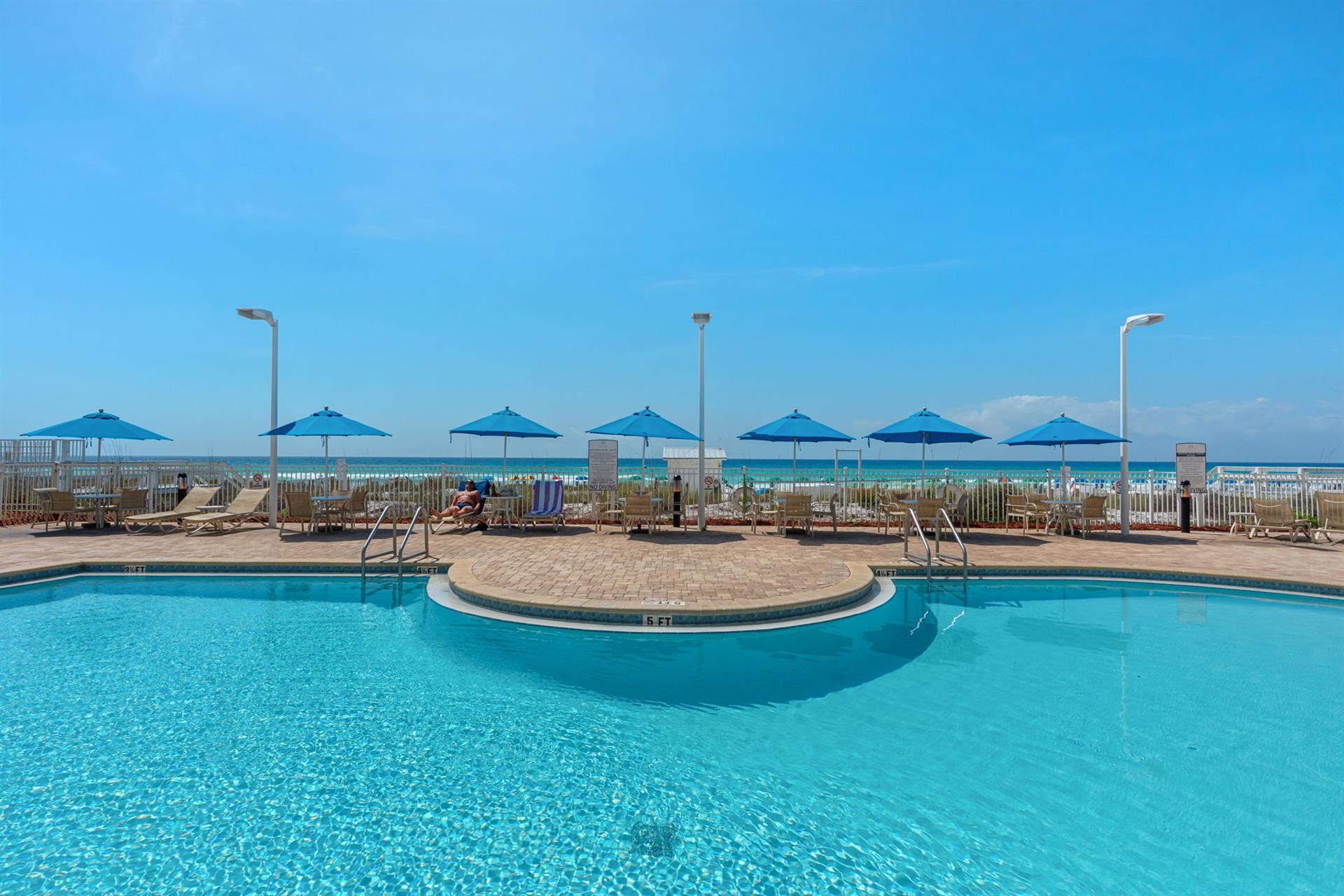 Sparkling pool with beachfront access featuring turquoise umbrellas and lounge chairs overlooking pristine sandy shores.