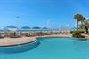 Beachfront pool area with blue umbrellas, lounge chairs, and direct access to pristine sandy shores under clear Florida skies.