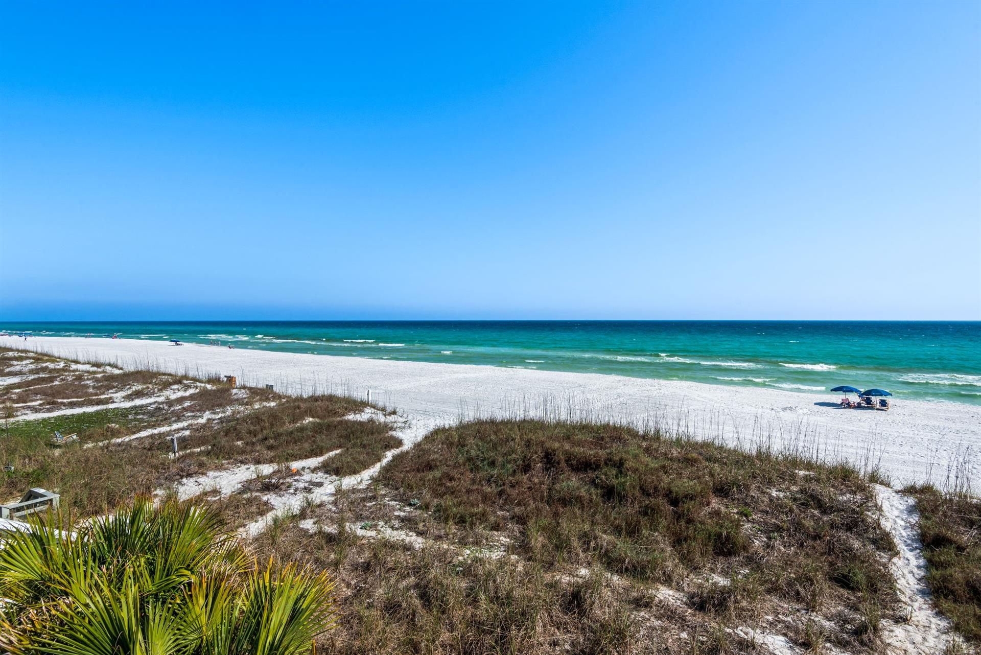 Pristine white sand beach stretches along emerald waters under clear blue skies, with natural dune vegetation and walking paths leading to the shore.