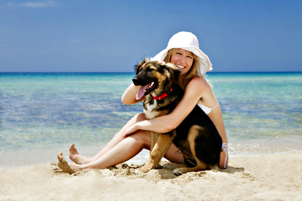 Blond woman sitting on sand hugging her dog on a dog-friendly beach.