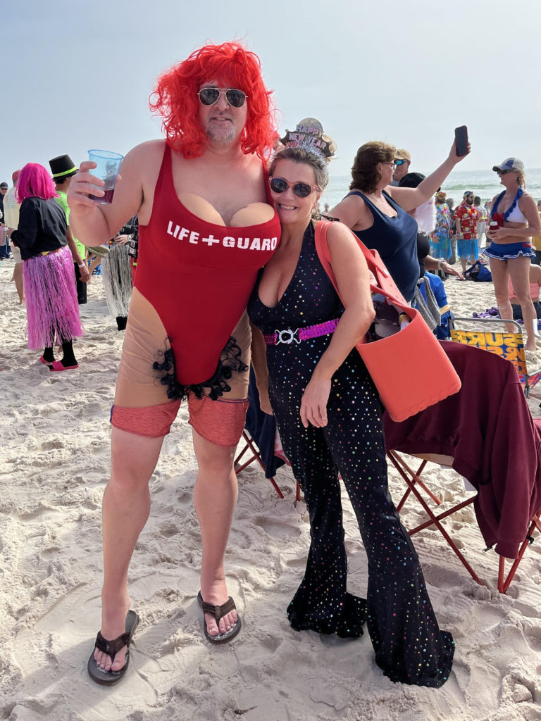 A man dressed as a woman in a one-piece read bathing suit that reads "Life+Guard" with fake breasts and hair  poses with a woman dressed in a disco jumpsuit on the beach during the 2023 Flora-Bama Polar Bear Dip.