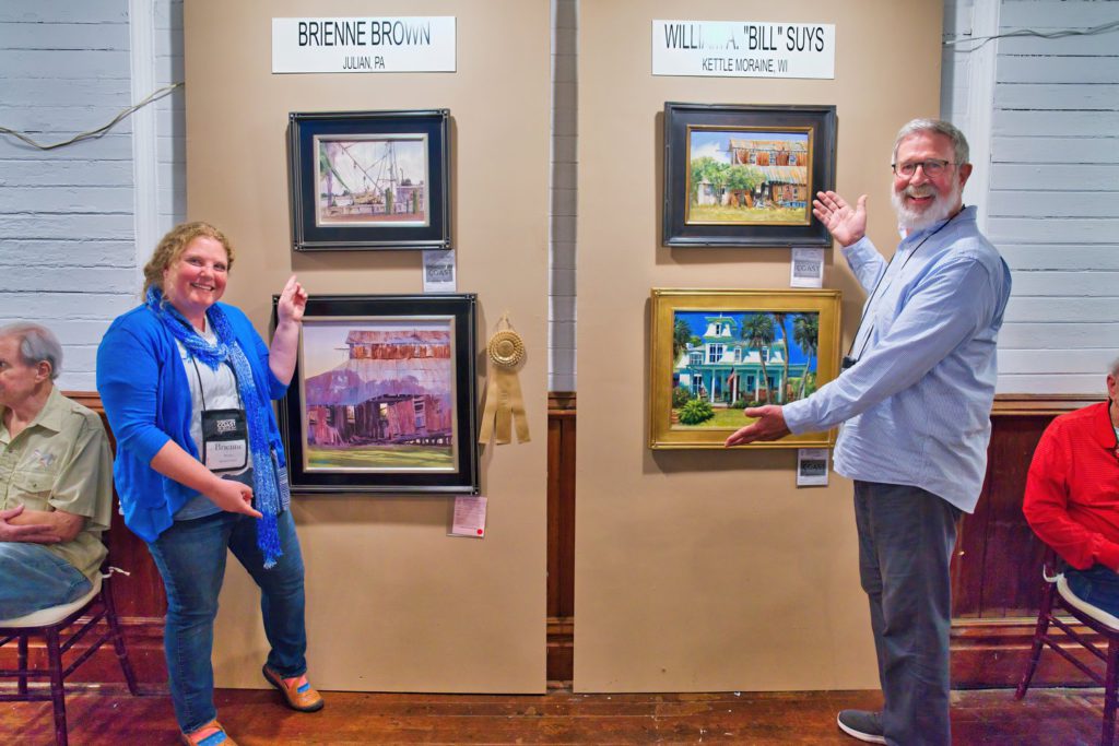 En Plein Air participants point to their framed paintings hanging on a wall.