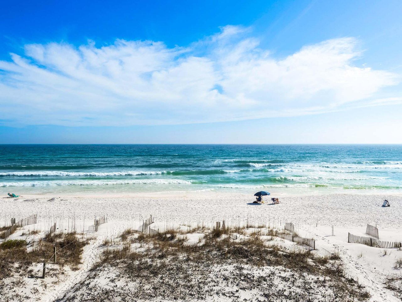 Pristine white sand beach stretches along turquoise waters under dramatic cloudy skies, with gentle waves and scattered beachgoers enjoying the coastal paradise.