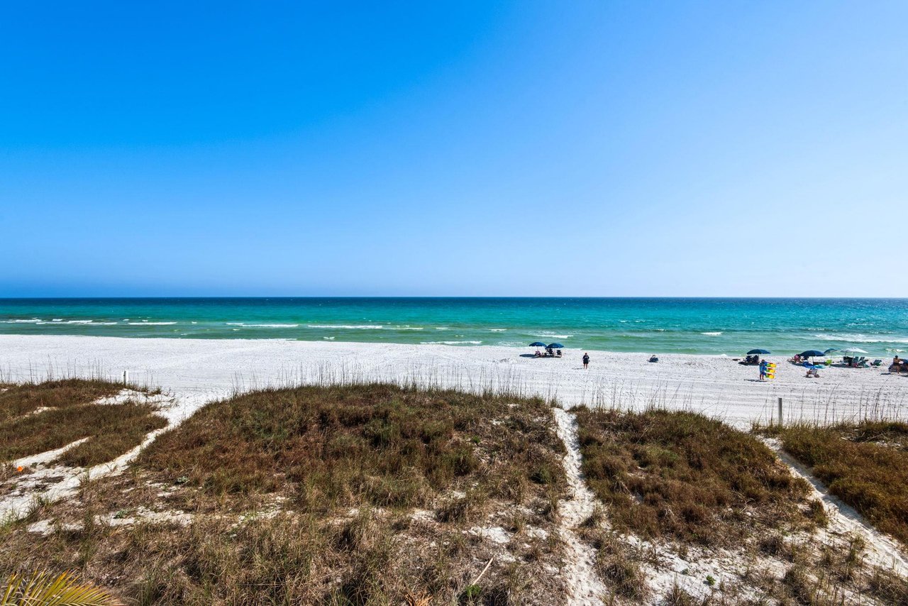 Pristine white sand beach stretches along turquoise waters under clear blue skies, with natural dune vegetation creating coastal pathways.