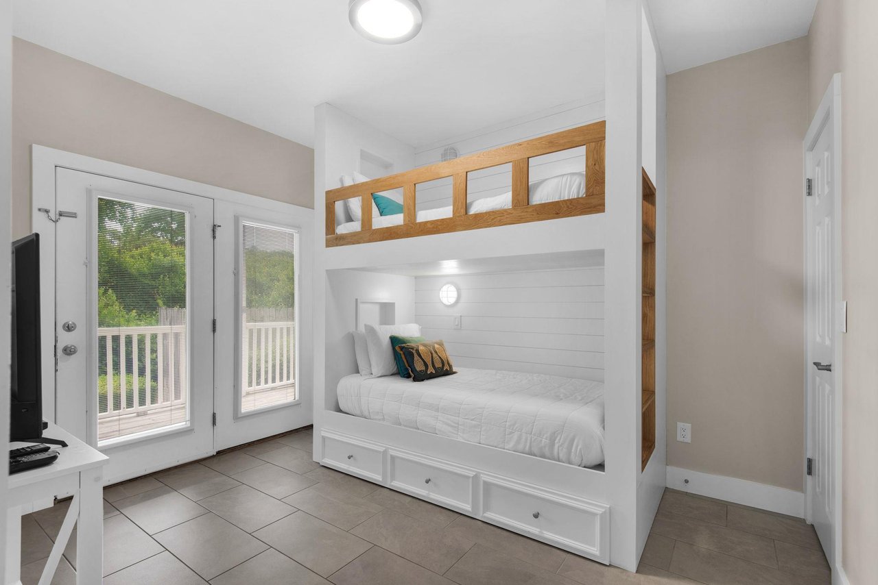 Bedroom 3 1st Fl features custom white shiplap beds with cozy storage drawers, natural light streaming through doors to the deck, and all the restful charm you need after a beach day.
