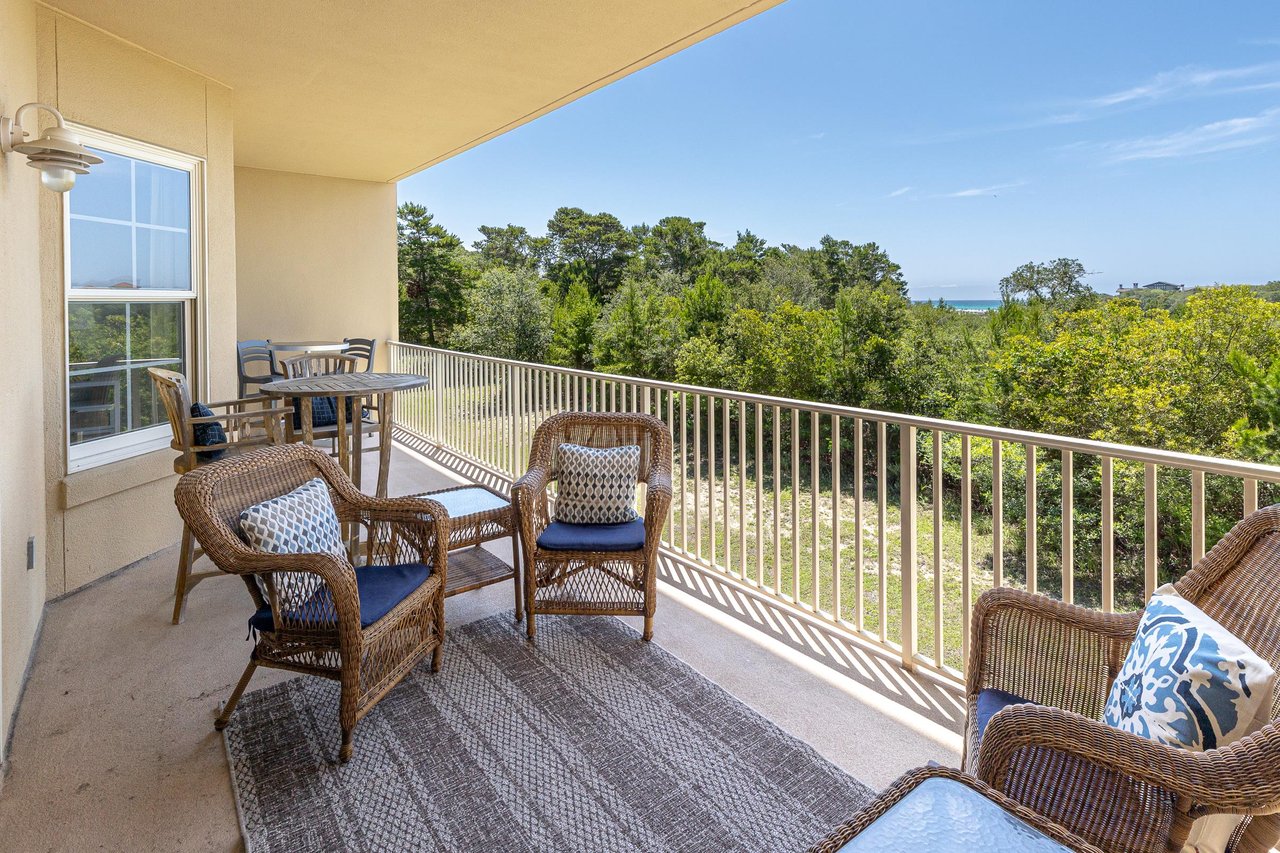 Expansive balcony overlooking lush treetops and distant sea views, featuring wicker seating with blue cushions and a dining table—perfect for morning coffee or sunset relaxation.
