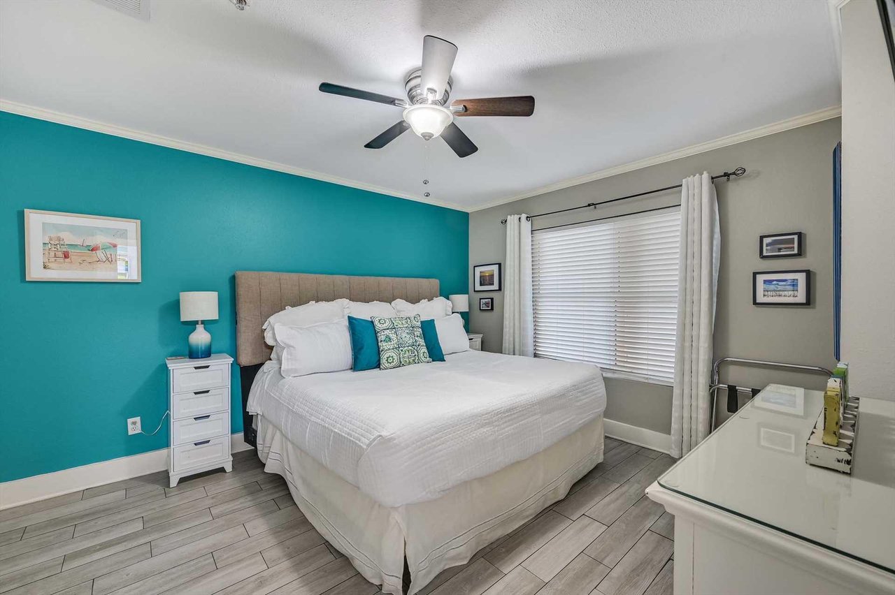 Step into Bedroom 2 providing bright, coastalinspired bedroom with a plush bed and soothing turquoise accent wall. The ceiling fan keeps you cool while natural light floods through the window.