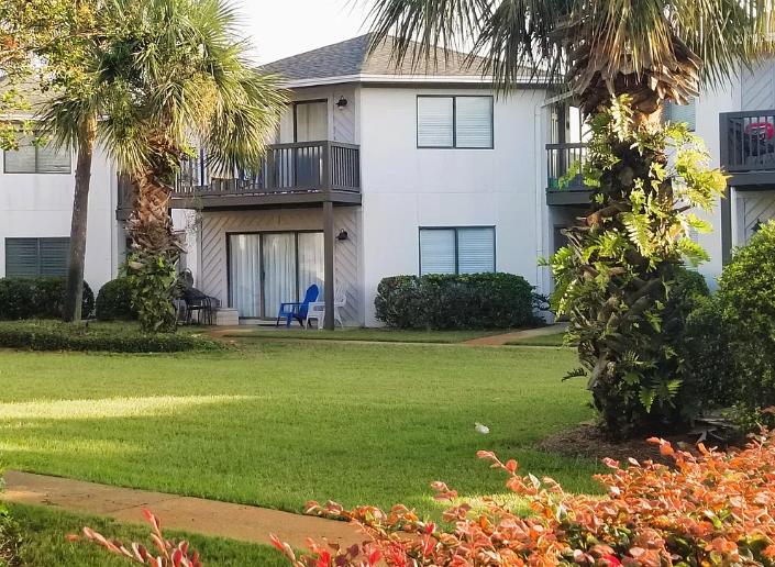 The property sits nestled among palm trees and tropical landscaping, with a groundfloor patio and an upper balcony overlooking vibrant greenery and lush lawn areas.