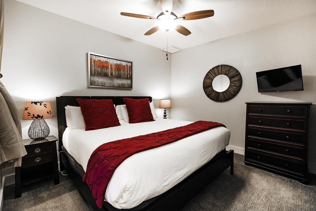 Guest bedroom with king bed, white linens and duvet and red accent runner and throw pillows, wall mounted TV and wooden dresser