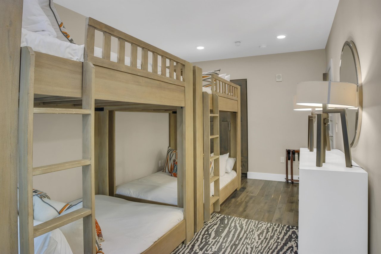 Two light colored wood bunkbeds, pushed end to end with a grey and white floor rug and wood floors.