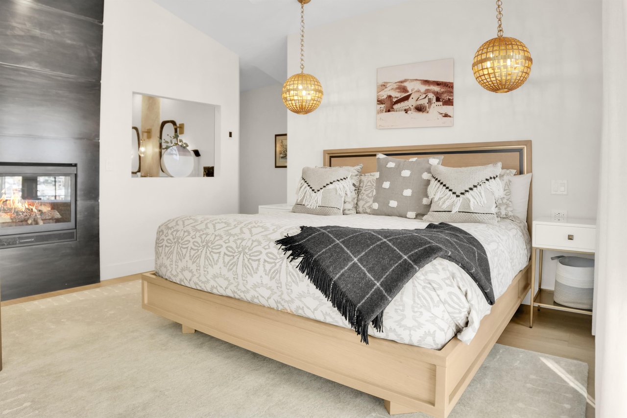 The light colored wood kingsized bed has grey and white pattened bedspread and throw pillows, and a dark grew throw blanket.