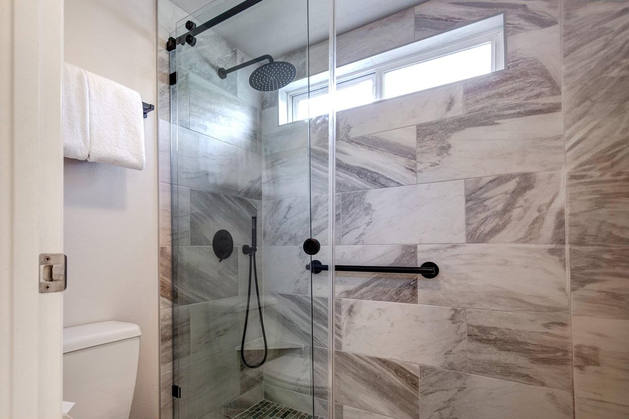 The walkin shower has grey and white patterned wall tile, black hardware, white toilet, and a white folded hanging towel.
