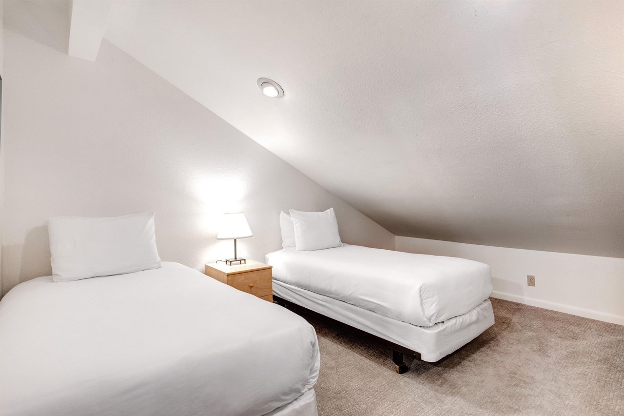 2 twin beds with crisp white linens, shared night stand with a lamp, and a pitched ceiling with recessed lighting.