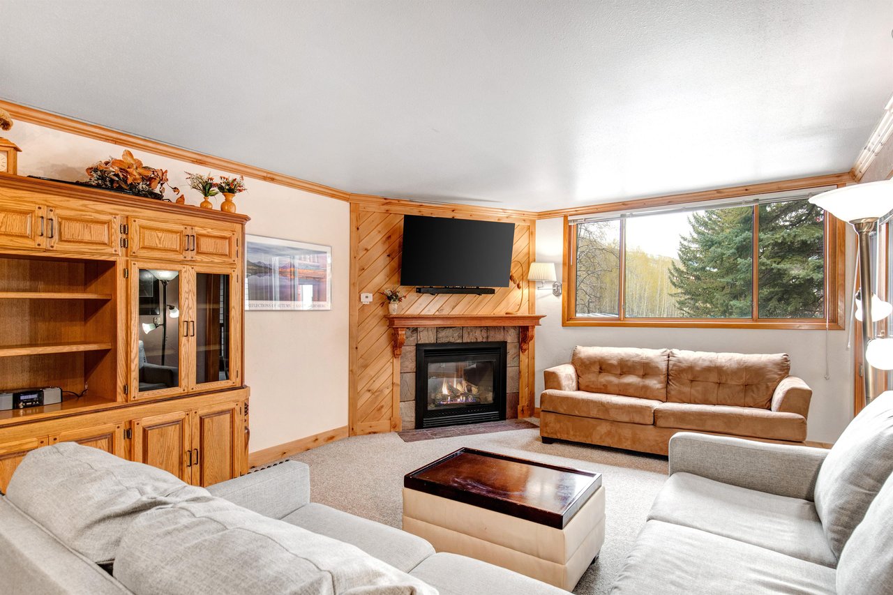 Snow Flower 43s living room has a large window framing a mountain and forest, grey sectional, leather couch, and fireplace.