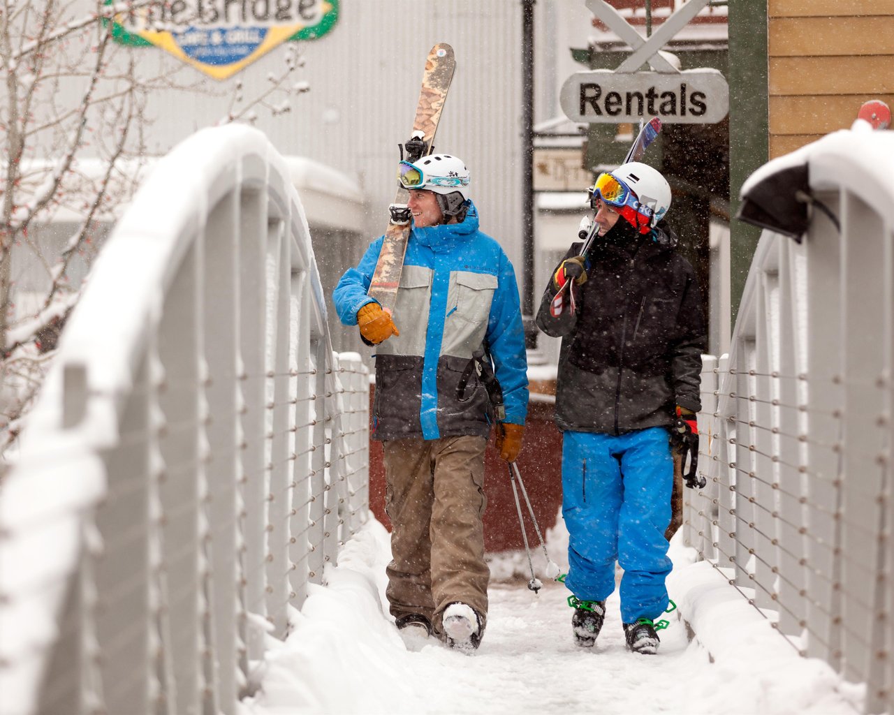 Two skiers walking their skis across the skier access bridge from their skiinskiout property.