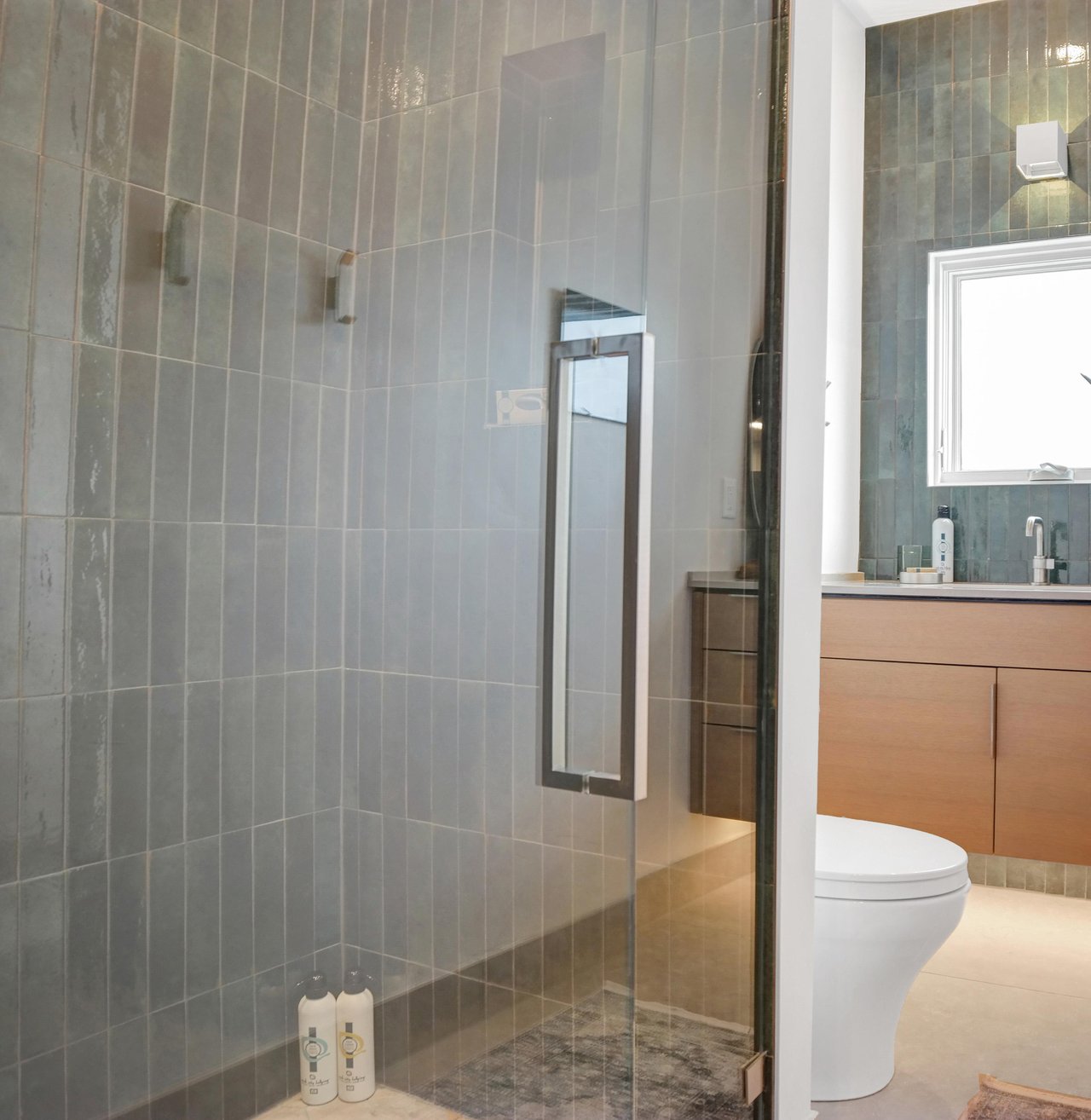 The bathroom has a walk in shower with deep green tile surround and glass doors and wood vanity with deep green tile backsplash.