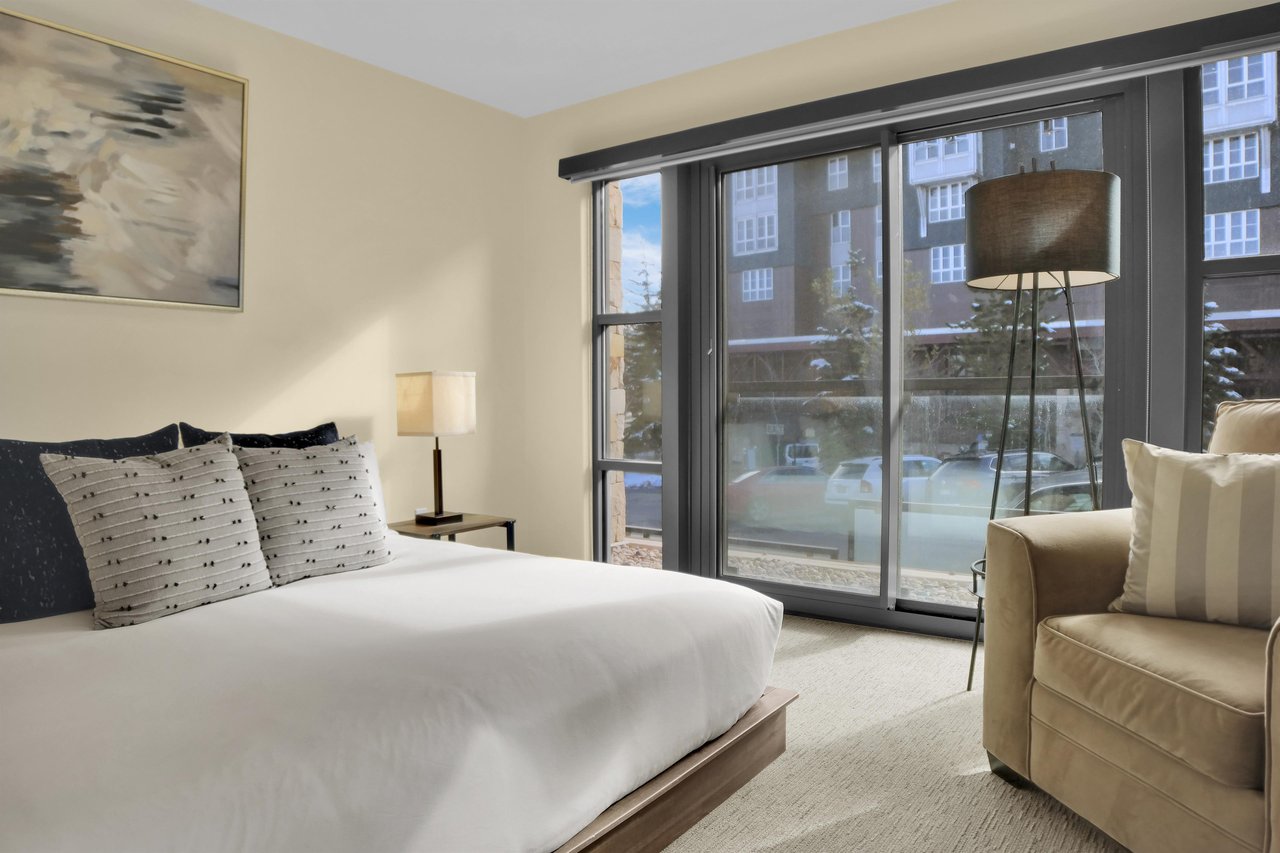 A king bed with crisp white linens, end tables with lamps, grey throw pillows, tan chair, floor lamp, and large windows.