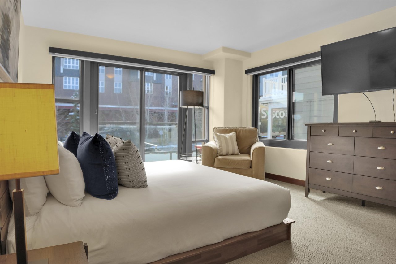 A king bed with crisp white linens, grey throw pillows, tan chair, floor lamp, wood dresser and flat screen TV.